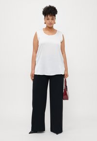 Donna con capelli ricci, che indossa una blusa bianca senza maniche, pantaloni neri a gamba larga, scarpe nere con punta, orecchini d'oro, mentre tiene in mano una piccola borsa rossa.