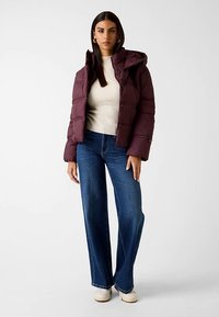 Kurze lila Steppjacke mit Kapuze, Druckknopfverschluss vorne und Seitentaschen; kombiniert mit einem hellfarbenen Pullover und weit geschnittenen blauen Jeans.