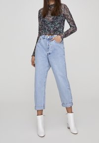 Top lungo a maniche trasparenti con fantasia floreale e collo alto, abbinato a jeans blu chiaro a vita alta con risvolti alle maniche e stivali bianchi alla caviglia.