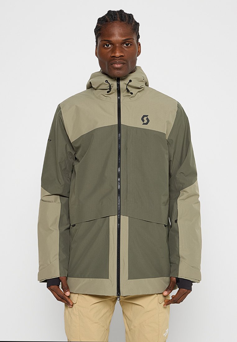 Chaqueta exterior verde y beige con cierre de cremallera, capucha ajustable y logotipo en el pecho. Presenta un diseño bicolor y una tela texturizada.