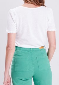 Blusa blanca texturizada con mangas cortas abullonadas y puños con bordes festoneados, combinada con pantalones verdes de cintura alta con grandes bolsillos traseros.