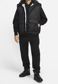 Gilet imbottito nero con zip frontale, due tasche frontali e interno bianco, abbinato a un felpa nera e pantaloni della tuta coordinati. Sneakers nere.