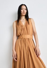 Robe sans manches, décolletée en V, de couleur beige chaud, avec des plis, des embellissements cloutés le long du décolleté, et une taille ceinturée.