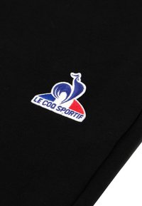 Svart tyg med en patch som har en blå, vit och röd logotyp samt texten "LE COQ SPORTIF." Patcher är broderad med en len yta.
