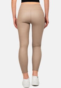 Donna vestita con leggings aderenti beige e sneakers bianche, in piedi con le mani ai fianchi su uno sfondo neutro.