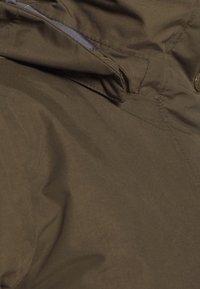 Brun, lätt jacka med hög krage, synlig sömnad och en slät textur. Har en knäppning med knapper och innertyg i grått.