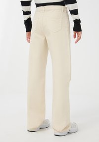 Pantaloni a gamba larga in denim di colore crema, con vita alta, due tasche posteriori e un orlo leggermente sfrangiato su un lato.