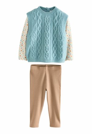 Gilet lavorato a maglia azzurro chiaro con motivi a treccia, indossato sopra una camicia crema con pois colorati, abbinato a leggings beige a coste.