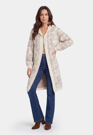 Kvinde iført lang beige mønstret cardigan, blå jeans, brune støvler og farverige øreringe, stående med den ene hånd på hoften.