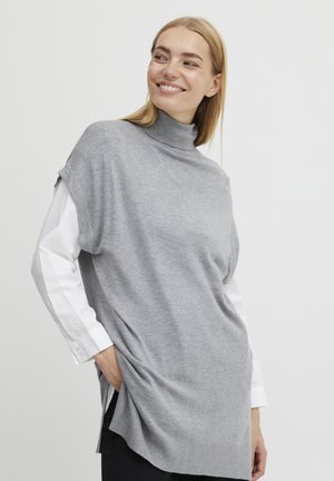 Grijze gebreide trui met een hoge hals, korte mouwen en zijsplitten, gelaagd over een witte lange mouwen shirt met knopen bij de manchetten.