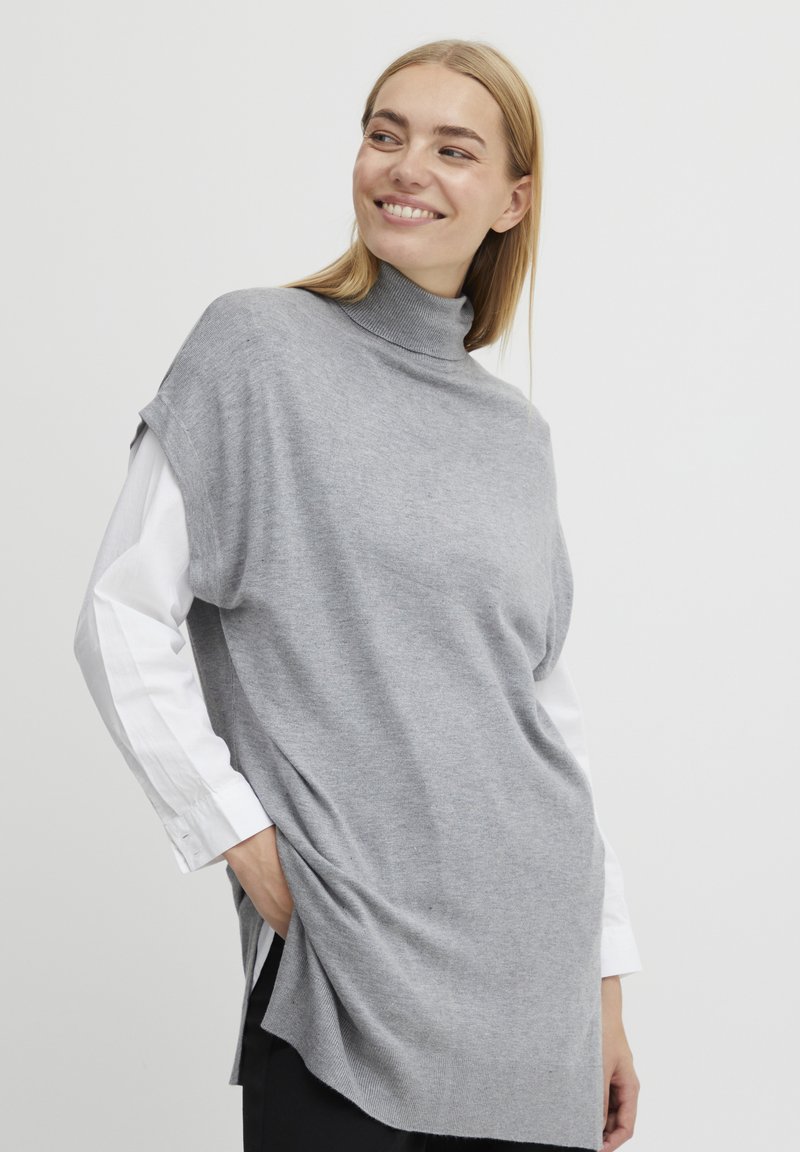 Grijze gebreide trui met een hoge hals, korte mouwen en zijsplitten, gelaagd over een witte lange mouwen shirt met knopen bij de manchetten.
