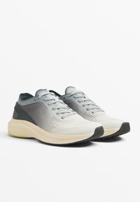 Világosszürke atlétikai cipők texturált hálós felsőrésszel, sötétszürke díszítésekkel és párnázott krémszínű talppal. Fűzőkkel és lekerekített orr-résszel rendelkeznek.