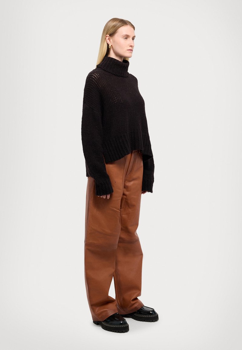 Schwarzer, gestrickter Rollkragenpullover mit lockerer Passform, kombiniert mit braunen, ledernen Schlaghosen und klobigen schwarzen Schuhen.