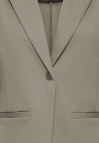 Blazer taupe à un bouton avec revers crantés et deux poches à passepoil sur le devant, sur un fond neutre.