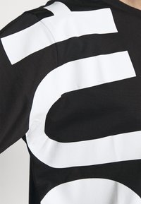 Sort langærmet skjorte med fed, oversized hvid grafik design, der indeholder tykke vandrette og buede former hen over skuldrene og brystet.