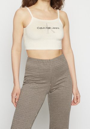 Nainen pukeutuneena vaaleaan Calvin Klein Jeans -lyhytpaitaan ja korkeavyötäröisiin ruskeavalkoruudullisiin housuihin yksiväristä taustaa vasten.