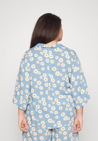 Top azul com padrão de margaridas brancas e amarelas. Apresenta gola, mangas curtas e um corte solto. O tecido parece leve com uma textura suave.
