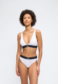 Γυναίκα με σγουρά μαλλιά που φοράει λευκό σουτιέν Calvin Klein και αντίστοιχα εσώρουχα με μαύρες ελαστικές ζώνες με το λογότυπο, στέκεται μπροστά σε απλό φόντο.