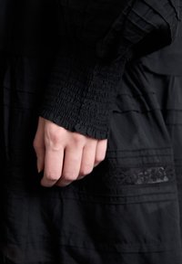 Črna blago obleka z teksturiranim gubanjem na rokavu. Diskretne vodoravne črte in čipkasta obroba na žepu. Površen posnetek roke, ki počiva.