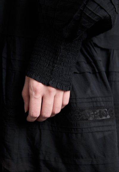 Zwart stoffen kledingstuk met gestructureerde smocking op de mouwen. Subtiele horizontale strepen en kantdetail op de zak. Close-up van een rustende hand.