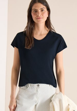 Kvinde med langt mørkt hår iført en sort kortærmet top og hvide bukser med striber, der holder en lysefarvet jakke i venstre hånd.