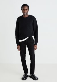 Musta neule, jossa on pyöreä kaula-aukko, valkoisen paidan päällä, yhdistettynä mustiin slim-fit-farkkuihin ja mustiin kenkiin, seisoo neutraalia taustaa vasten.