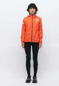 Veste imperméable orange vif avec fermeture éclair, associée à des leggings noirs moulants et des baskets noires, portée par un modèle en position debout.