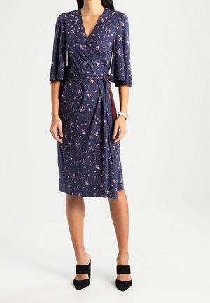 Kvinde iført navy blå knælængde slå-om kjole med små lyserøde blomsterprint, 3/4 ærmer og sorte mule med hæl.