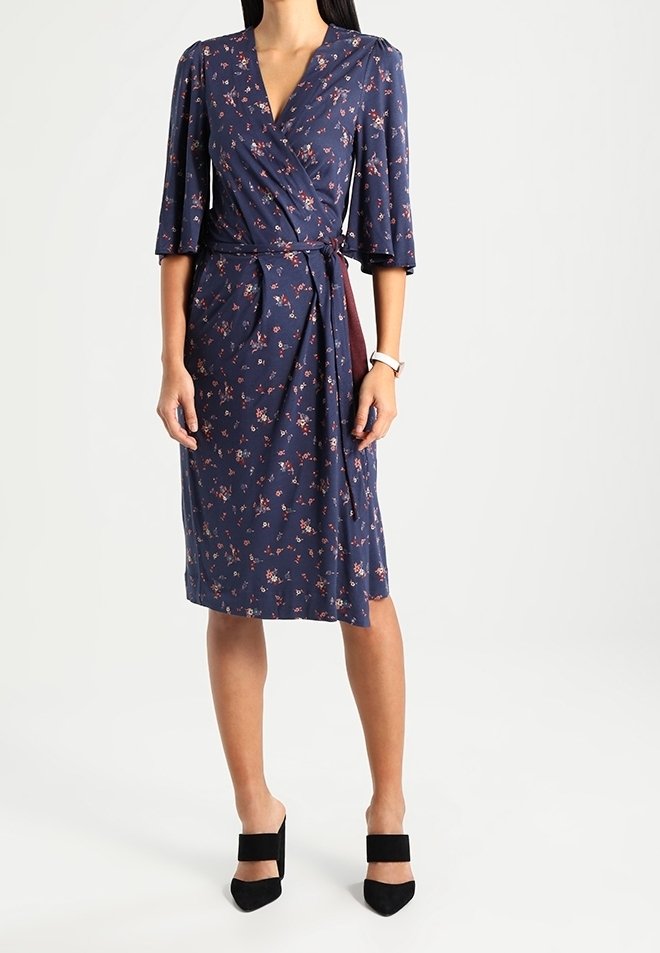 Femme portant une robe portefeuille bleu marine longueur genoux avec un petit imprimé floral rose, manches trois-quarts, et des mules noires à talons.