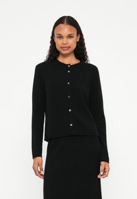 Μαύρο, μακρυμάνικο καρφίτσα φτιαγμένο από μαλακό ύφασμα με στρογγυλή λαιμόκοψη, διαθέτοντας ασημί κουμπιά κατά μήκος του μπροστινού μέρους. Απλός πλεκτός σχεδιασμός.