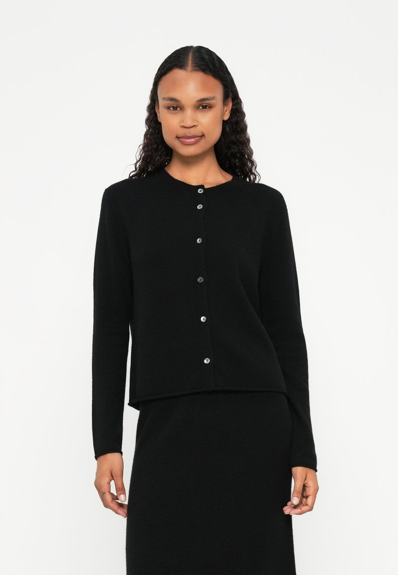 Μαύρο, μακρυμάνικο καρφίτσα φτιαγμένο από μαλακό ύφασμα με στρογγυλή λαιμόκοψη, διαθέτοντας ασημί κουμπιά κατά μήκος του μπροστινού μέρους. Απλός πλεκτός σχεδιασμός.