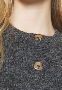 Tmavě šedý pletený oděv s texturovaným povrchem, zdobený dvěma kulatými světle hnědými knoflíky na výstřihu.