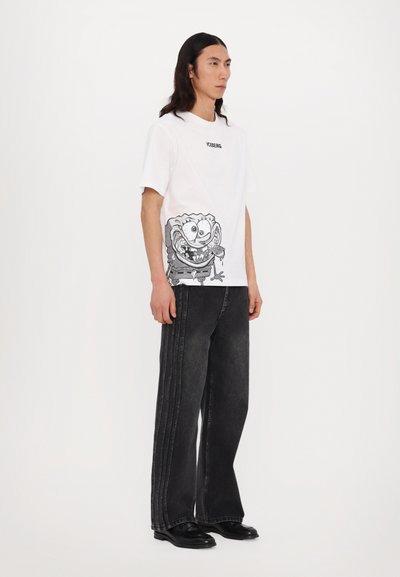 T-shirt blanc à manches courtes avec un design graphique d'un personnage de dessin animé en bas. Assorti à un jean large noir et des chaussures noires.