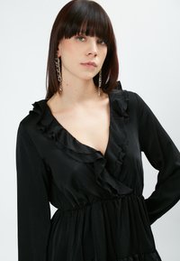 Vestido negro con escote en V con volantes, mangas largas y cintura fruncida. Confeccionado con una tela suave y fluida para una textura delicada.