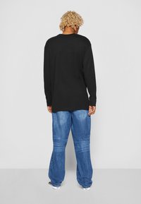 Chemise noire à manches longues associée à un pantalon en denim bleu ample. La chemise présente un col rond ; le pantalon a une texture délavée et des jambes larges.