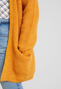 Närbild av en person som bär en bygig orange stickad cardigan över blåjeans och en mönstrad skjorta, med ena handen i cardiganens ficka.