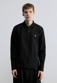 Μαύρο μακρυμάνικο πόλο μπλουζάκι με τρεις κουμπότρυπες, κατασκευασμένο από βαμβάκι. Διαθέτει μικρό ορθογώνιο λογότυπο της μάρκας στο στήθος.