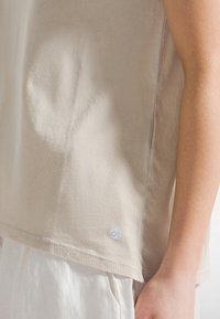 Beige bomulls-T-shirt med avslappnad passform, rundad nederkant och en liten logotyp-tag på sidan. Mjuk textur och slät yta.