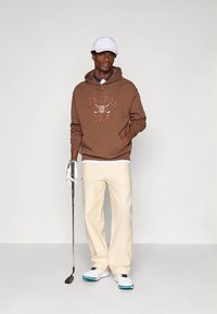 Pruun kapuutsiga dressipluus tekstiga "MALBON GOLF", kreemjad püksid, valge nokamüts ja valged golfijalatsid; hoiab golfikeppi, seisab neutraalse taustaga ees.