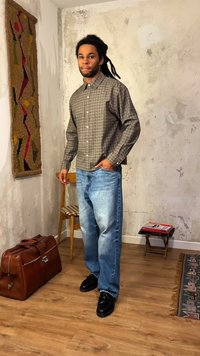 Άνδρας με dreadlocks, ντυμένος με καφέ πουκάμισο καρό, μπλε τζιν και μαύρα παπούτσια, στέκεται σε ένα ρουστίκ δωμάτιο με δερμάτινη τσάντα και ξύλινη καρέκλα.