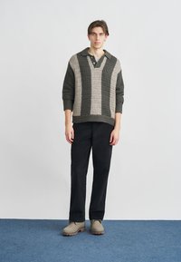 Strikkegenser i mørkegrønn og beige, med V-hals, vertikale striper og et teksturert mønster. Kombinert med svarte bukser og beige sko.