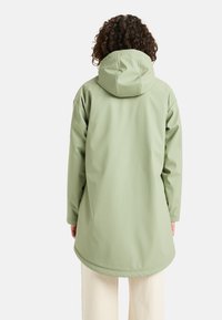 Svetlozelený vodeodolný kabát s kapucňou, s voľným strihom, dlhými rukávmi a zaobleným lemom, vyrobený z hladkého, matného materiálu.