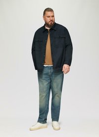 Donkere marineblauwe knoopjas over een bruine trui, gecombineerd met lichtblauwe denim jeans en witte sneakers. Model staat tegen een effen achtergrond.