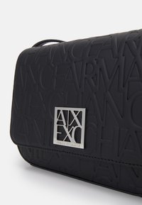 Schwarze Umhängetasche aus gummiertem Material mit erhabenen Logo-Mustern und einem silbernen Hardware-Akzent. Kompakte rechteckige Form.
