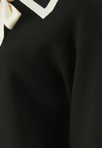 Tissu noir avec un col blanc présentant un design en V et un détail de nœud. Texture douce et lisse avec une apparence épurée et ajustée.
