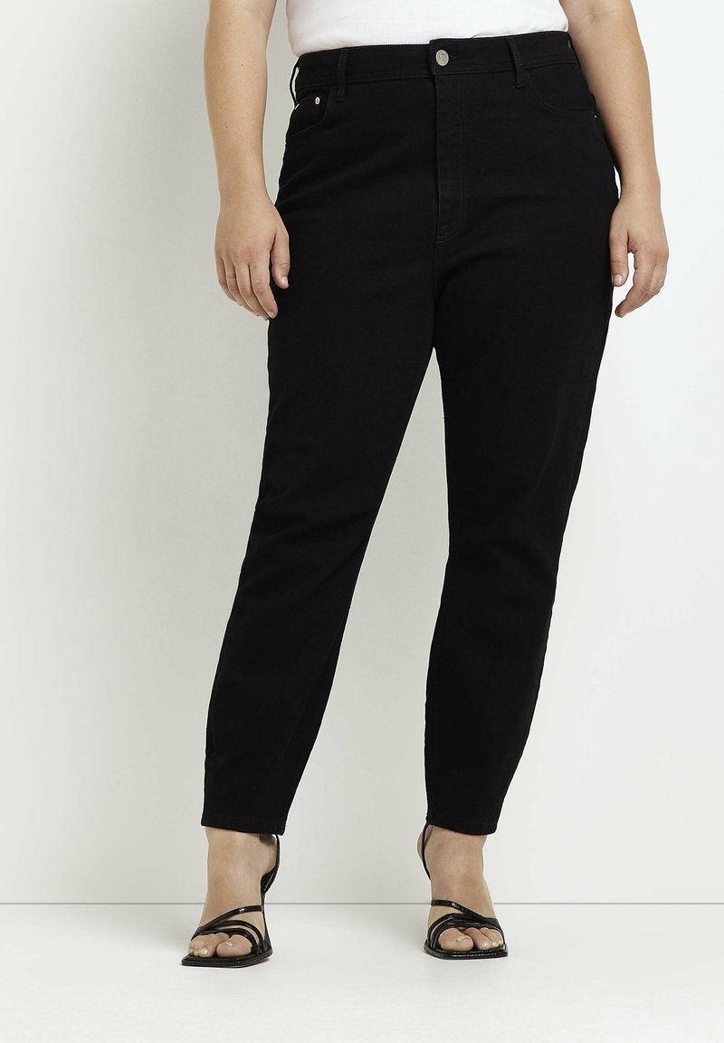 Personne portant un jean noir taille haute et des sandales noires ouvertes, debout devant un fond clair uni.