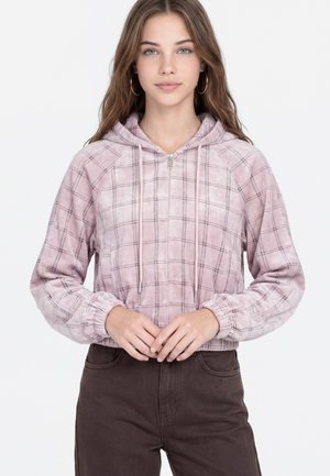 Young woman with long hair wearing hoop earrings, a pink plaid zip-up hoodie, and dark pants, standing with hands clasped.