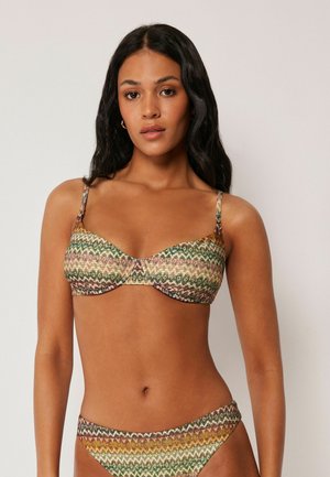 Kobieta o długich ciemnych włosach, ubrana w wzorzysty zielono-brązowo-żółty bikini, stojąca na tle jednolitego jasnego tła.