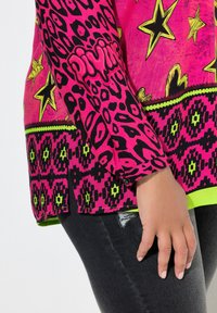 Hand rustend naast zwarte gescheurde jeans, gekleed in een levendig roze langetermijntop met gele sterren en een zwart luipaardpatroon.