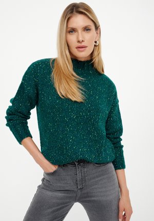 Groene gebreide trui met hoge hals, gestructureerd patroon en lange mouwen, gecombineerd met grijze jeans.